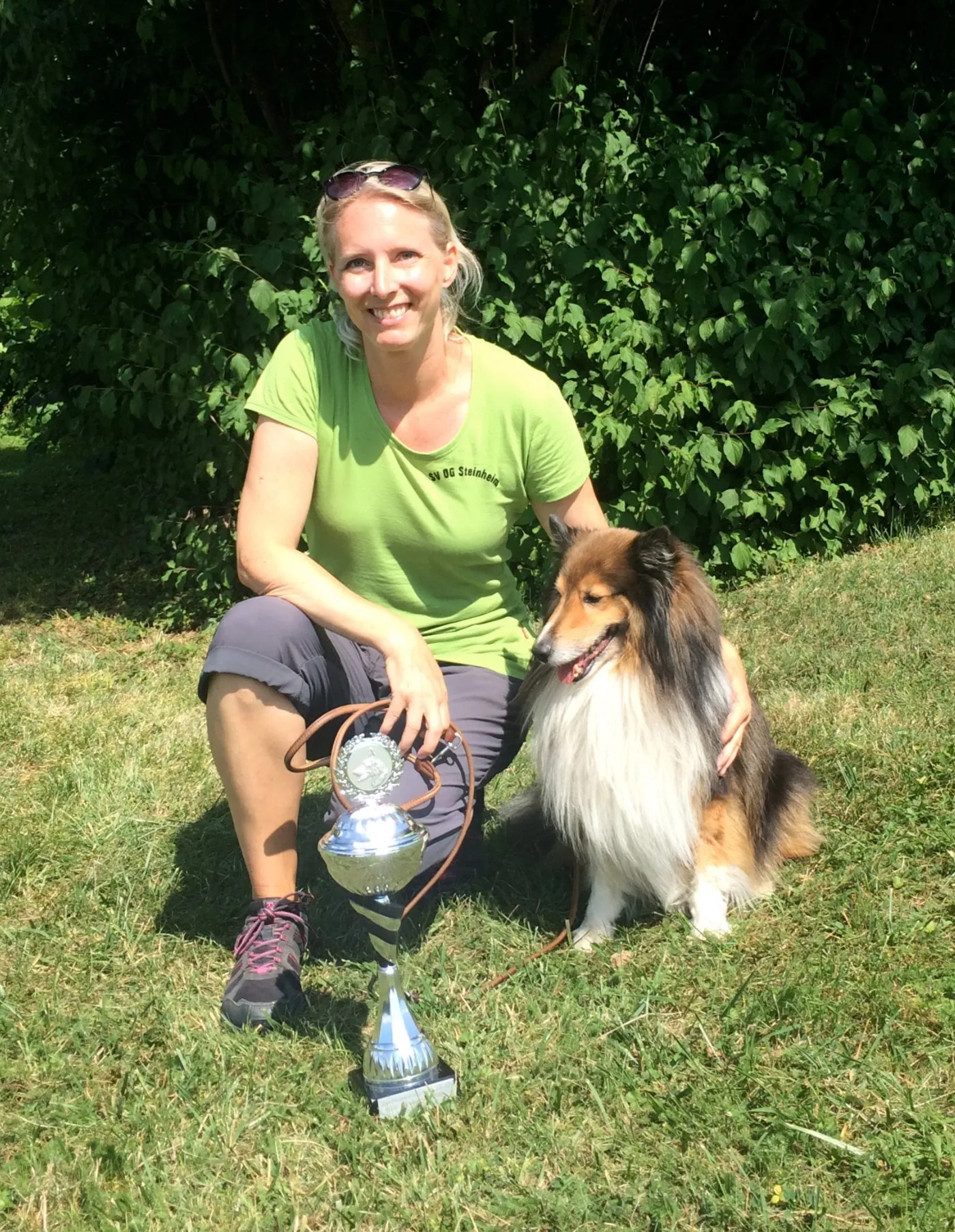 Jana Elzenbeck kniet lächelnd im Gras neben ihrem braun-weißen Sheltie Nosy, vor den beiden steht ein silberner Pokal